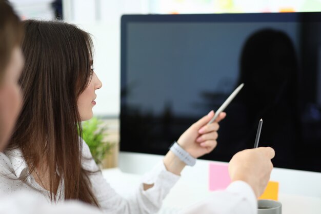 Portrait of smart businessman and businesswoman sitting in modern workplace and pointing at high-tech computer screen with pens