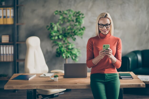 Portrait of smart company owner woman use smartphone chatting with employees colleagues stand near wooden table in office loft wear red turtlneck