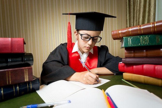 Ritratto di ragazza intelligente in cappello di laurea che fa i compiti in biblioteca