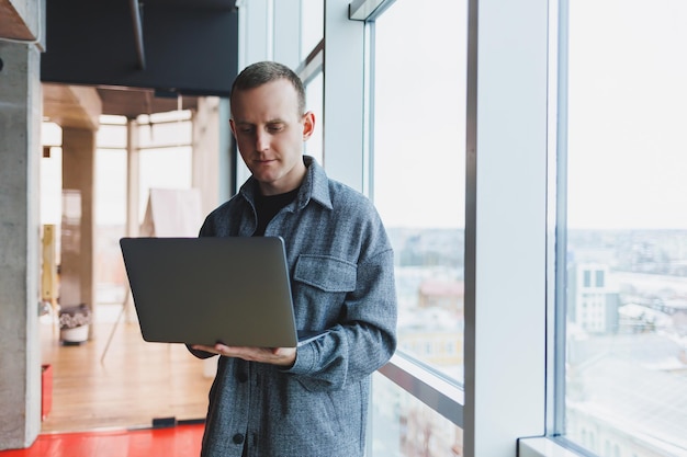 사무실에서 연구를 위해 노트북을 들고 있는 똑똑한 남성 관리자의 초상화는 우아한 옷을 입고 터치패드에서 노는 사업가 큰 창 옆에 사무실에 서 있습니다.