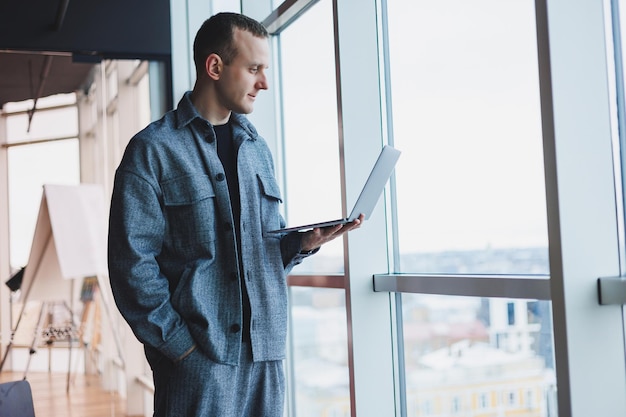 Портрет умного интеллигентного менеджера-мужчины, он держит ноутбук для исследований в офисе, деловой человек в элегантной одежде, играющий на тачпаде, стоящий в офисе у большого окна