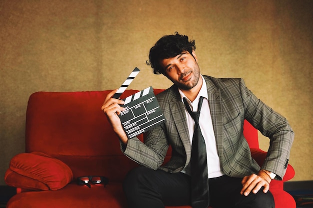 Portrait smart man sit on red sofa holding movie clapboard.