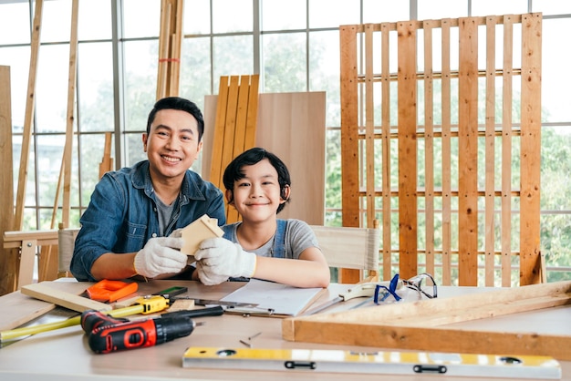 Ritratto di sorriso asian padre e figlio falegnameria seduta costruendo e tenendo una piccola casa di legno insieme a casa studio di falegnameria con lo strumento di misura e strumenti di lavoro sul tavolo