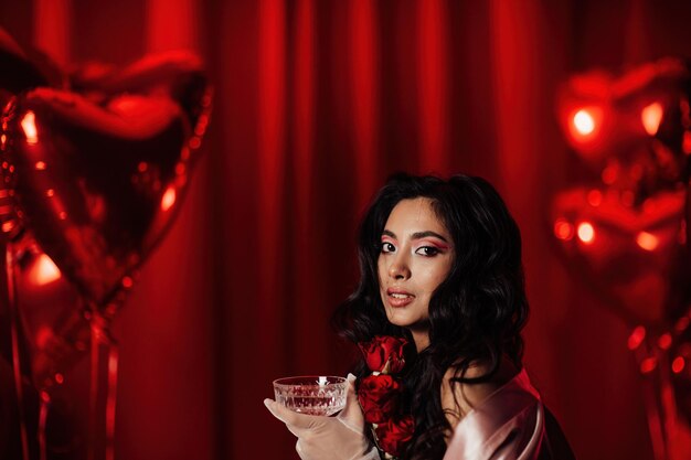 Портрет улыбающейся сексуальной азиатской девушки в гламурном макияже в нижнем белье с красными розами и бокалом шампанского купе
