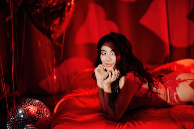 Портрет улыбающейся сексуальной азиатской девушки с гламурным макияжем в красном кружевном белье, лежащей на кровати в день святого валентина