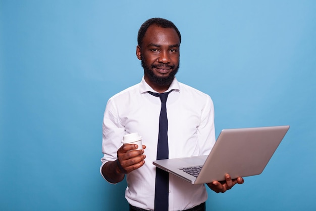 Portrait of smiling accountant looking at camera holding laptop and cup of hot to go drink on blue background. Businessman casually using wireless portable computer while having a coffee.