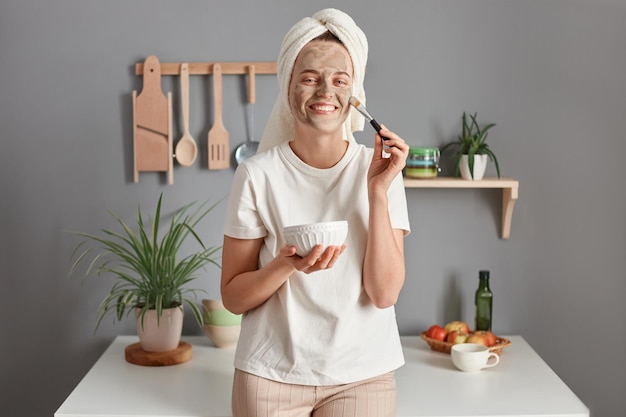 Photo portrait of smiling adorable woman with bath towel on her head applying anti aging facial mask to her face at modern kitchen holding cosmetic brush skin care treatment at home