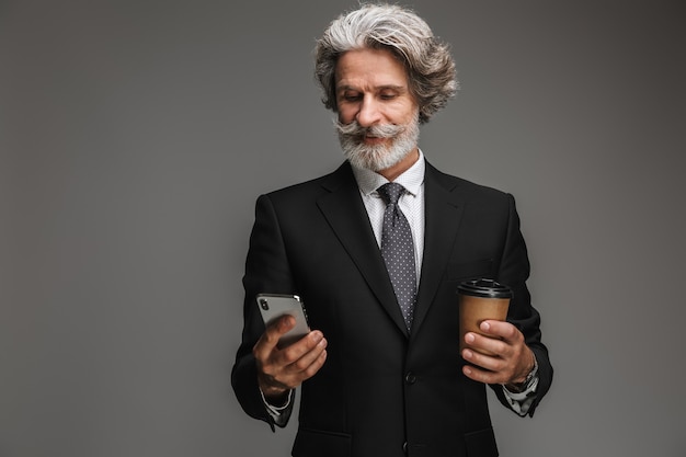 회색 벽에 격리된 종이컵과 스마트폰을 들고 공식적인 검은 양복을 입은 웃고 있는 성인 사업가의 초상화