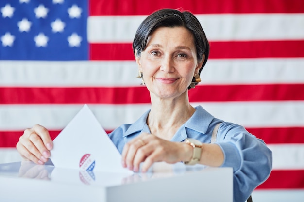 Ritratto di donna adulta sorridente che mette il voto nel cestino contro la bandiera americana