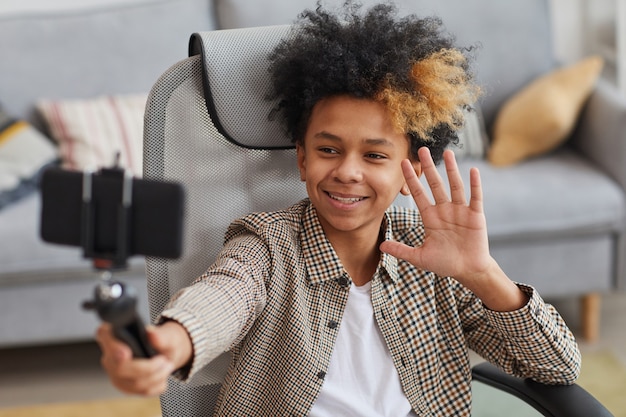 Portrait of smiling African-American boy waving at camera while holding selfie stick and video blogging from home, copy space