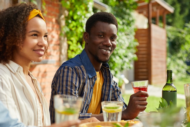 Портрет улыбающегося афроамериканца, наслаждающегося ужином с друзьями на открытом воздухе, сидя за столом во время летней вечеринки