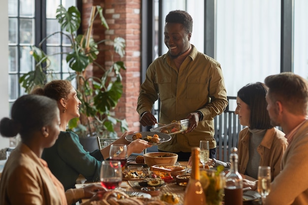 Portrait of smiling African-American man serving food while hosting dinner party with friends and family at home, 