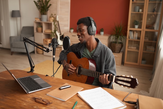 Portrait of smiling African-American man singing to microphone and playing guitar while recording music in studio, copy space