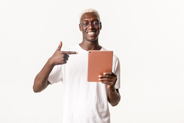 Portrait of smiling african-american stylish blond guy in glasses, reading e-book in digital tablet, pointing satisfied at gadget