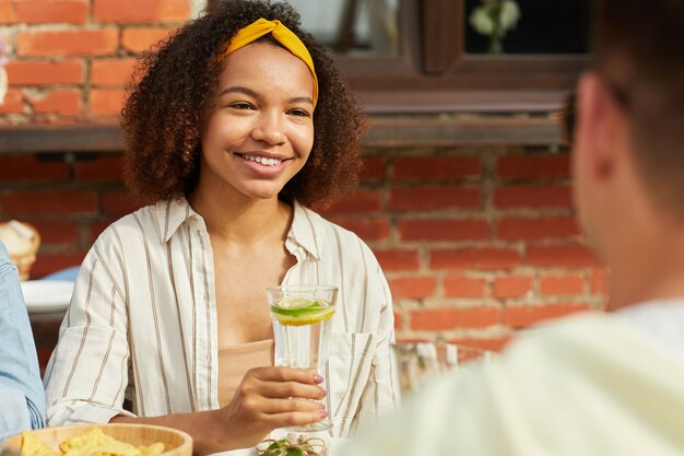 Photo portrait of smiling african-american woman enjoying dinner with friends outdoors and holding refreshing cocktail while sitting at table during summer party