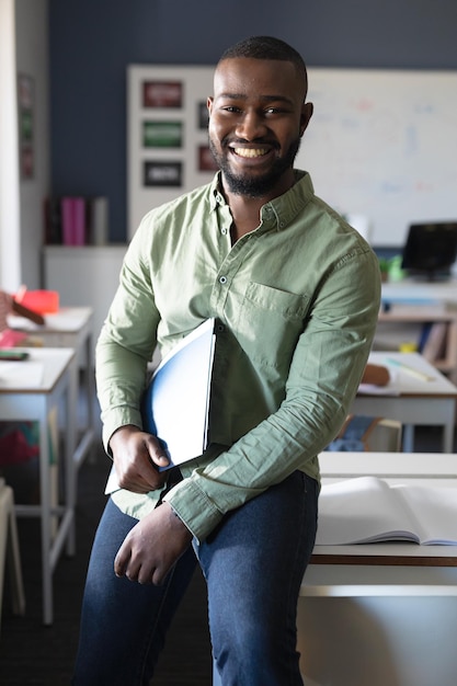 Portrait of smiling african american young male teacher with file sitting at desk in classroom