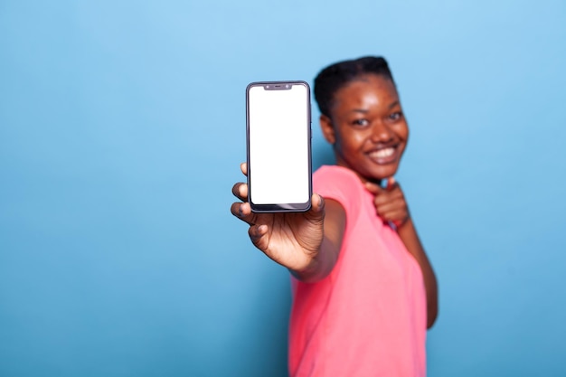 Portrait of smiling african american young woman showing smartphone with white display at camera standing in studio with blue background. Beautiful teenager holding phone with touchscreen.