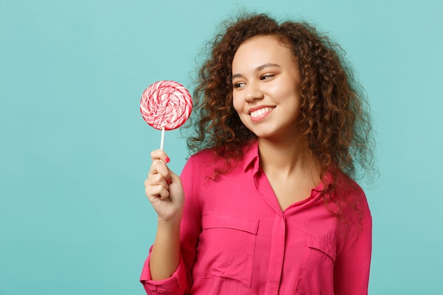 Portrait of smiling african girl in casual clothes hold, looking on pink round lollipop isolated on blue turquoise background in studio. People sincere emotions, lifestyle concept. Mock up copy space.