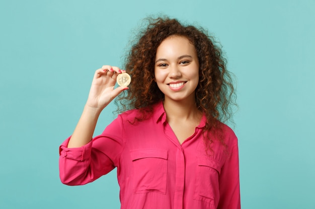 Portrait of smiling african girl in casual clothes holding bitcoin, future currency isolated on blue turquoise wall background in studio. People sincere emotions lifestyle concept. Mock up copy space.