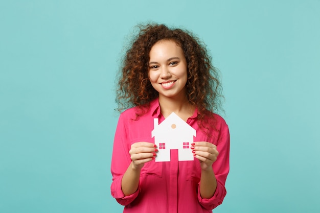 Ritratto di una ragazza africana sorridente in abiti casual rosa che tiene in mano una casa di carta isolata sul fondo della parete turchese blu in studio. concetto di stile di vita di emozioni sincere della gente. mock up copia spazio.