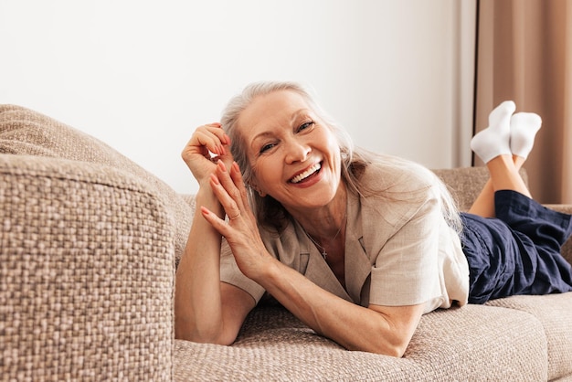 カメラを見てリビング ルームのソファに横たわっている白髪の笑顔の年配の女性の肖像画 x9xA