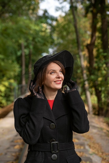 Portrait of smiling aristocratic woman in black hat with veil in autumn park Middleaged woman in vintage retro style