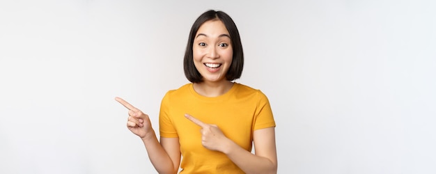 Ritratto di sorridente ragazza bruna asiatica in maglietta gialla che punta il dito a sinistra mostrando copia spazio promo affare dimostrando banner in piedi su sfondo bianco