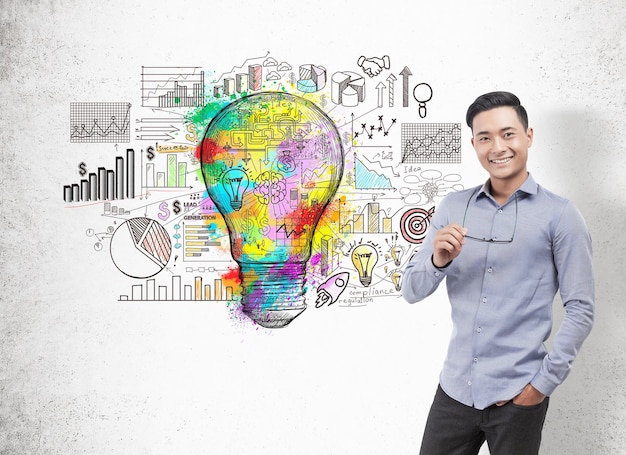 Portrait of a smiling Asian businessman wearing a blue shirt and dark trousers and holding glasses. Concrete wall background with a large colorful light bulb and a business scheme.