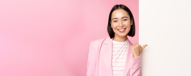 Portrait of smiling asian businesswoman in suit corporate lady pointing finger at white empty wall board with info or advertisement standing over pink background