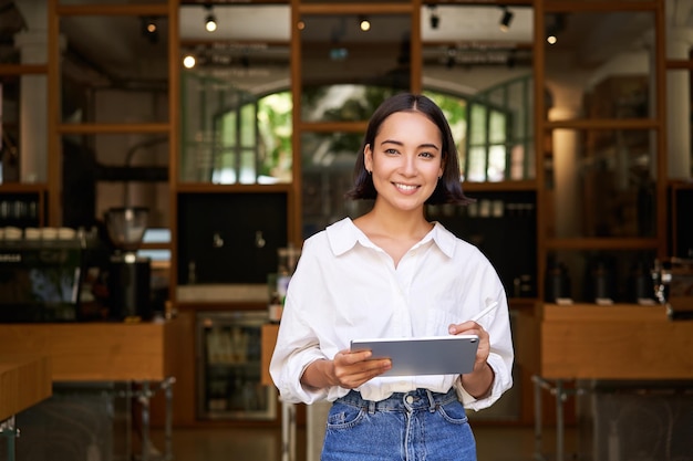 Портрет улыбающегося азиатского менеджера по персоналу кафе, стоящего перед входом с цифровым планшетом