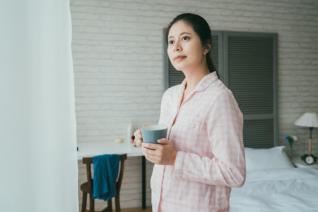 パジャマを着た笑顔のアジア人女性のポートレートが窓の近くに立ち、寝室で起きた後、晴れた朝とお茶を楽しみながら遠くを見ている。