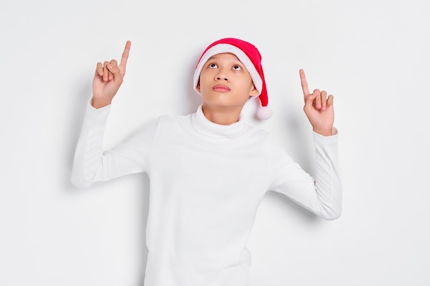 Portrait of smiling Asian man in christmas hat pointing fingers up inviting people to follow isolated on white background