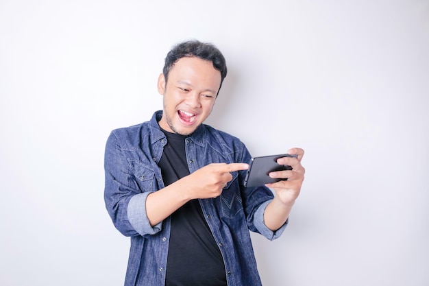 A portrait of a smiling Asian man is smiling and holding his smartphone wearing a navy blue shirt isolated by a white background