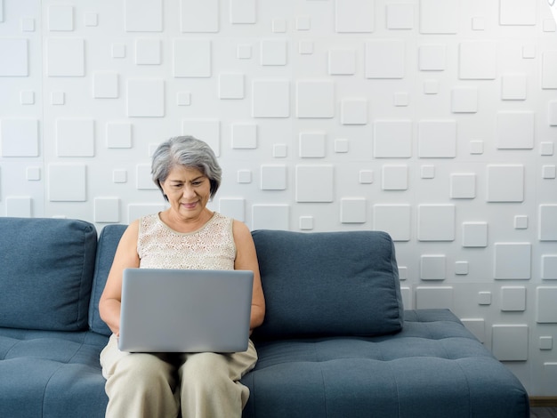 Портрет улыбающейся азиатской пожилой случайной женщины, сидящей на диване и работающей с ноутбуком в ярко-белой комнате Пожилая женщина занимается интернет-серфингом или видеозвонком дома Пожилые люди с технологиями