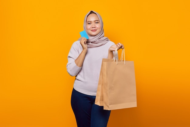 Portrait of smiling asian woman holding shopping bag and and showing credit card over yellow background