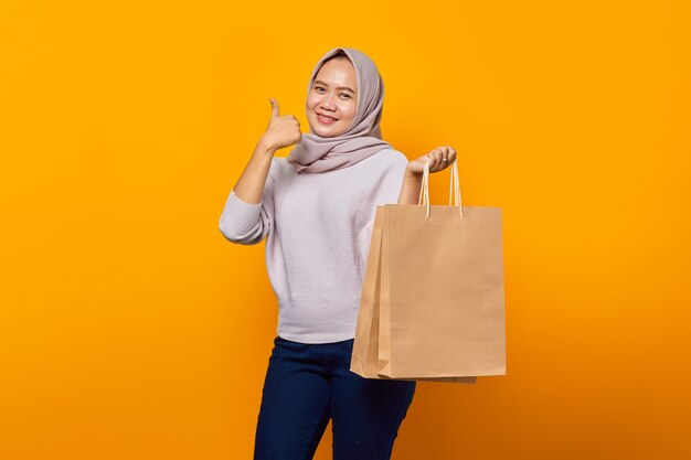 Portrait of smiling asian woman holding shopping bag and showing thumbs up over yellow background