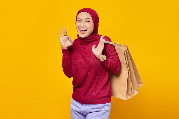 Portrait of smiling Asian woman holding shopping bags and making okay sign with fingers over yellow background