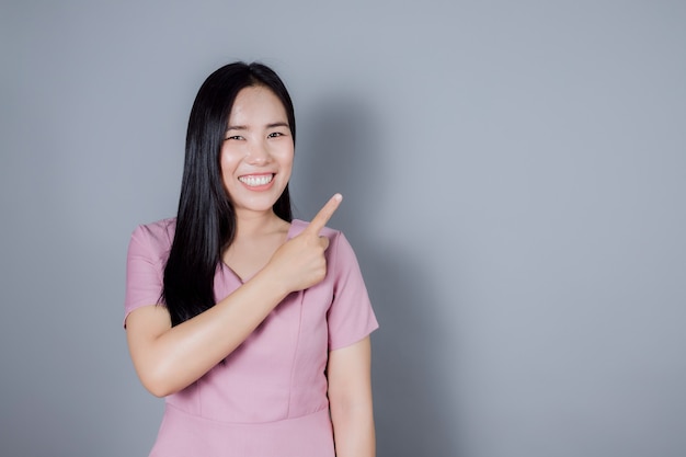 Ritratto di donna asiatica sorridente che punta il dito di lato su sfondo grigio