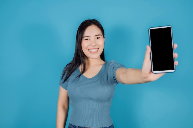 Portrait of smiling Asian woman showing blank screen mobile phone or smart phone on blue background