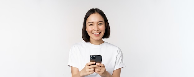 Portrait of smiling asian woman using mobile phone chatting texting message standing in tshirt over white background