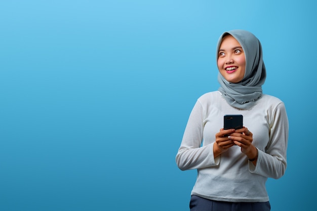 Portrait of smiling Asian woman using mobile phone and looking aside on blue background