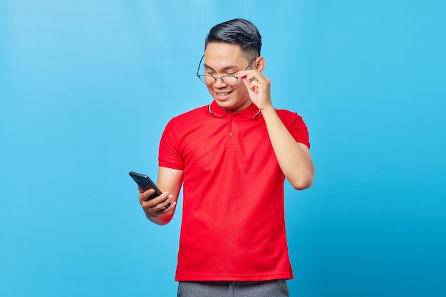 Portrait of smiling asian young man holding glasses and reading incoming message on mobile phone isolated on blue background