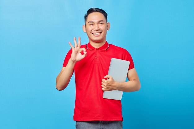 Portrait of smiling Asian young man in red shirt holding laptop and making okay gesture, showing approval symbol isolated on blue background