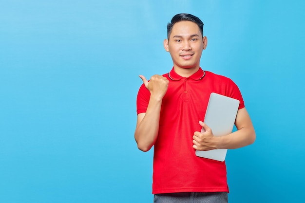 Portrait of smiling Asian young man in red shirt holding laptop and  pointing thumb up at copy space isolated on blue background