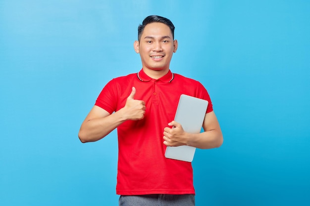 Portrait of smiling Asian young man in red shirt holding laptop and pointing thumbs up while looking at camera isolated on blue background