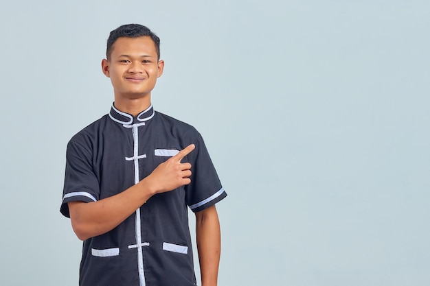 Portrait of smiling Asian young man wearing karate uniform and pointing sideways on gray background