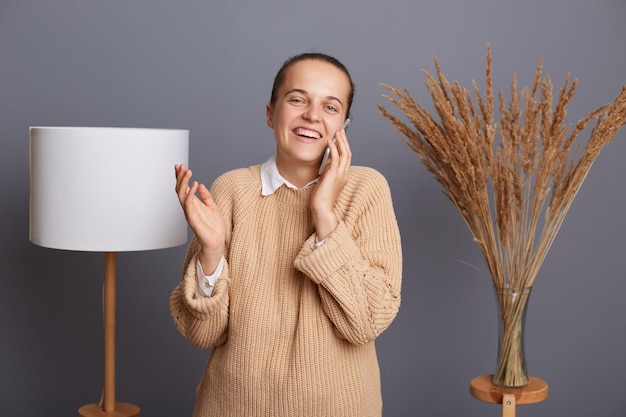 Портрет улыбающейся привлекательной позитивной женщины в теплом свитере, стоящей у серой стены с лампой и сушеным цветком на заднем плане, разговаривающей по мобильному телефону и выражающей счастье