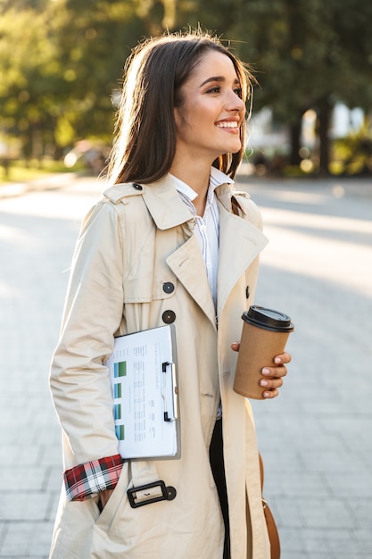 Portrait of smiling attractive woman wearing coat drinking takeaway coffee and holding clipboard while walking on city street