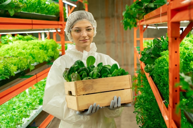 Portrait of smiling attractive young agroengineer in safety goggles and cap standing with box of plants at vertical farm