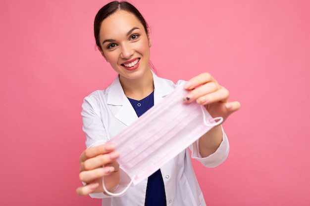 Portrait of a smiling attractive young female doctor in white coat holding medical mask standing